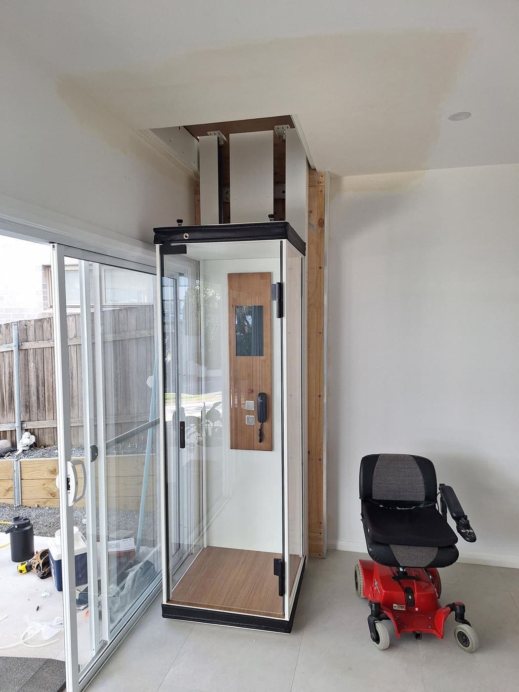 Residential through-floor home lift with glass doors, wired beside a mobility chair